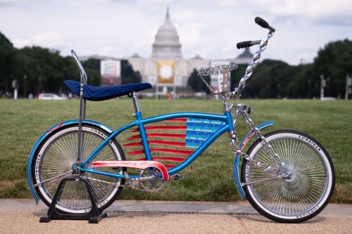 Shiny blue lowrider bike frame with red stripes and blue stars forming the American flag in the center of the frame. It is parked on the edge of the grass of the National Mall, with the U.S. Capitol Building in the background.