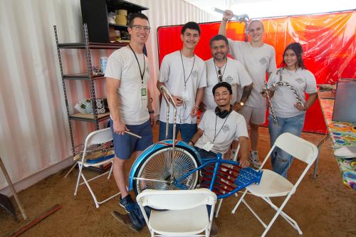Six people in matching white T-shirts sit and stand around the sparkly blue frame of a lowrider bicycle with decorative details to form the American flag. Some hold tools and pieces of chrome detailing.