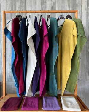 A rainbow of silk tunics hung on a wooden rack, in turquoise, bright pink, yellow, green, white, and crimson. Below them, four scarves in white and shades of purple folded on a shelf.