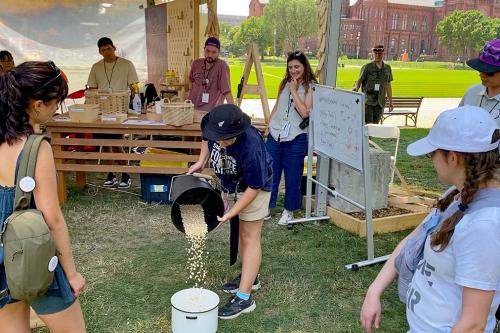 A young person pours kernels of white corn from one bucket into another on the ground, while others stand around and watch. In the background, a table is set with woven baskets and supplies.