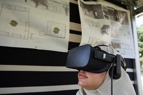 A person in a beige hijab wears a black Oculus virtual reality headset, looking up into the distance. Behind her, architectural plans for a courtyard building.