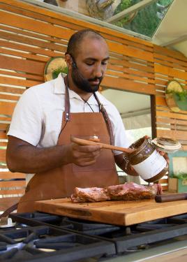 A man in a leather chef's apron spoons a dark brown wet spice mix onto a piece of a meat on a wooden cutting board.