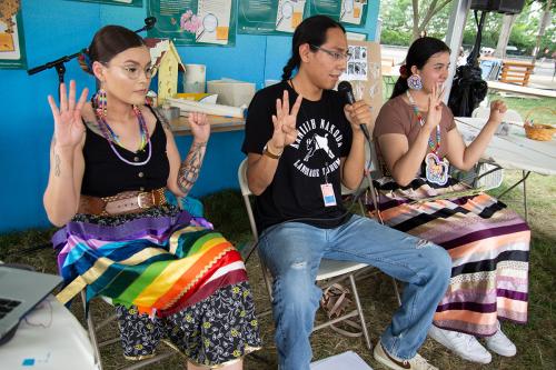 Three individuals are seated in chairs at an outdoor presentation. They each are holding their hands up, their left hand closed in a fist and the right hand with four fingers held up and the thumb down. The person in the middle is holding a microphone wit