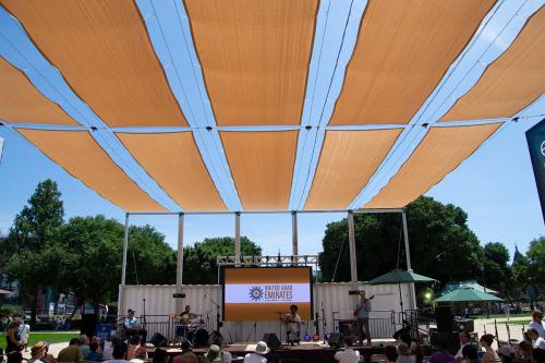 A three-piece band plays on an outdoor stage for a crowd, tan sun-shade panels stretched above. Behind them on stage, a screen reads United Arab Emirates.