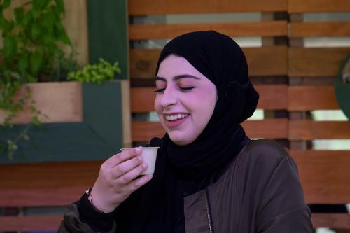A woman in black hijab holds a small paper cup, smiling.
