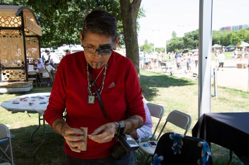 A woman with tribal tattoos on her eye and chin, and others on her forearms, sands a piece of stone under a tent outdoors, with crowds of people passing by in the background.
