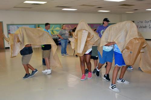 Pairs of people wear simplified, tan-colored buffalo costumes, which cover their heads and torsos, in a large room. 