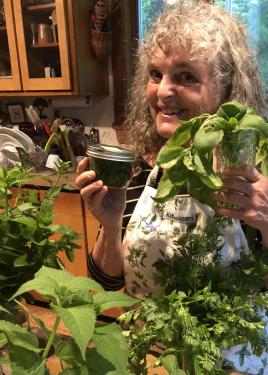 Susan, with gray wavy hair, holds up a sprig of green basil in a glass and a jar of green pesto. On the kitchen counter in front of her, more green herbs.