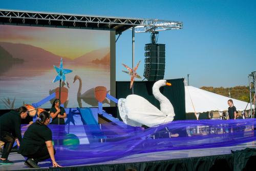 A white swan glides across stage. In the foreground, people dress in all black raise long pieces of blue translucent fabric, so it flows like water.