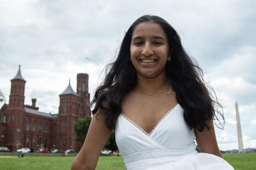 A young woman with a white dress and long black hair, smiling broadly, sits in the grass of the National Mall, with the Smithsonian Castle and Washington Monument in the background.