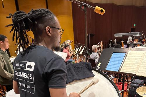 A young man plays timpani drums, reading sheet music from a stand. Beyond him are other players in an orchestra.