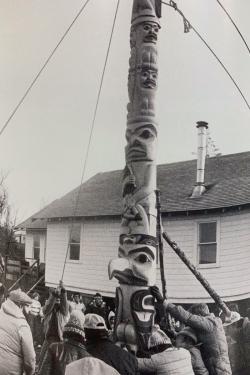 A crowd of people work together to raise a carved wooden totem pole outdoors with rope attached to the top. Black-and-white photo.