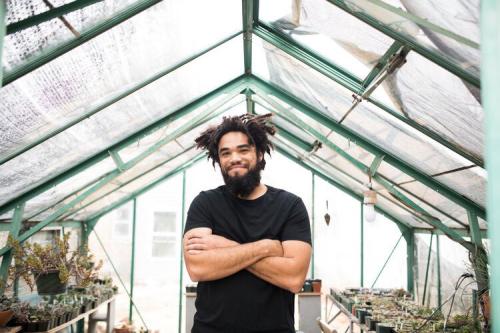 A Black with dark dreads and beard smiles, arms crossed, posing among plants in a bright greenhouse.
