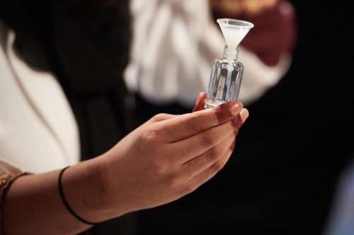 A person with henna-tipped fingers holds a small glass vial with a white funnel. Only their hands are visible.