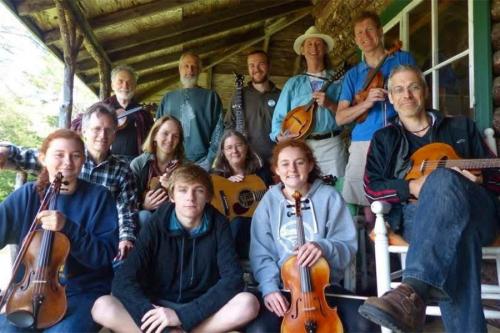 A group of twelve people, varying ages, sit in three rows and pose with their string instruments, including violins and guitars. 