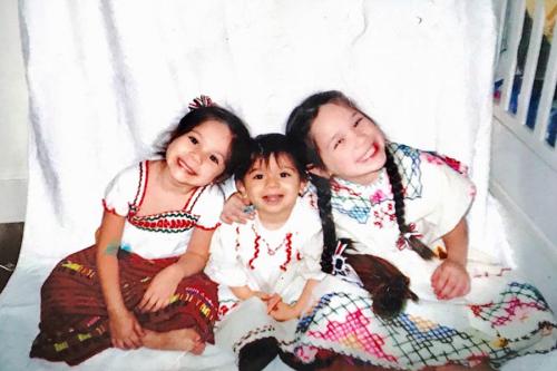 Two young girls sit and lean their heads on a young boy; the three of them sit on a white sheet and wear traditional Mexican regalia.