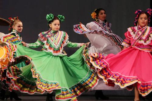 Four girls synchronously dance on stage, wearing different colors of the same Mexican dance dress.