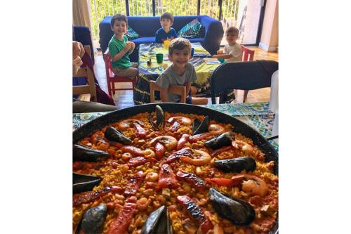 Four young boys sit around a kids' table turn around to smile at a person holding a pan of shrimp and oyster paella.