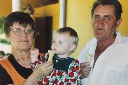 An adult woman holds a blonde baby while feeding her an icecream cone, the icecream is all over the baby's mouth; the two are joined by an adult man who also holds a melting icecream cone.