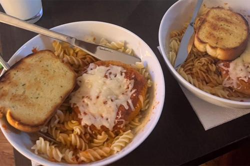 Two white bowls filled with pasta and bread covered in tomato sauce, cheese, and garlic sit on a dark wooden table.