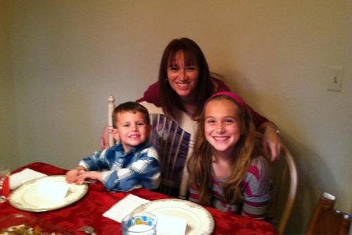 A woman, young girl, and younger boy smile; the boy and girl are sitting at a a table and the woman has her arms over both of their chairs.