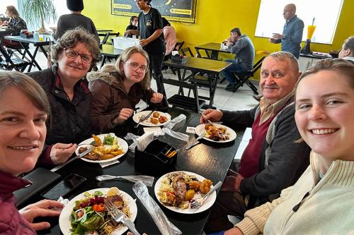 Four women and a man smile for a selfie while eating food at a restaurant table.