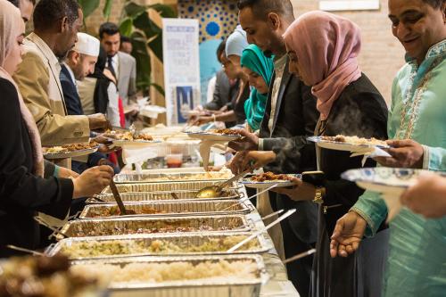 A crowd of people line up on both sides of a long buffet table, filling their plates with food. The women are wearing hijabs and the men suits or traditional tunics.