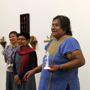 Three woman stand in an art gallery with white walls. The one on the right speaks as she holds a plain white Tree of Life figure.