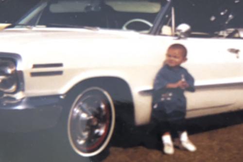 A toddler stands in front of a white lowrider, his head not even reaching the sideview mirror. Old color photograph.