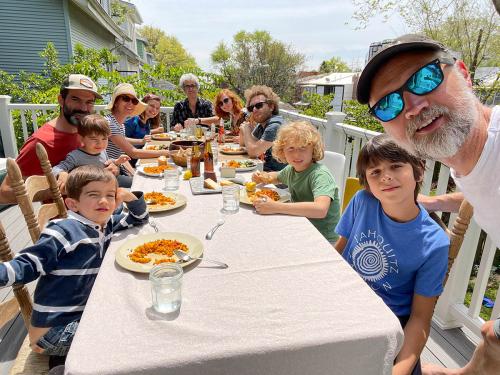 A group of eleven people, ranging from young children to adults, sit around a long table on an outdoor porch while eating paella.