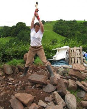 A young woman wields a hammer overhead with both hands, aimed at a slab of rock at her feet.