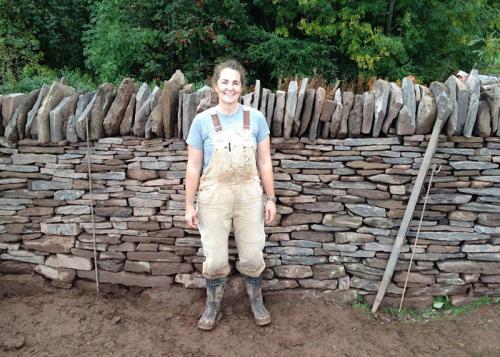 The woman, in dirty overalls and boots, poses in front of a stone wall with vertical slabs of stone placed decoratively along the top.