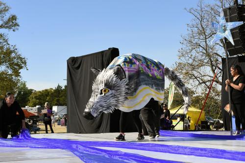 On stage, a gray wolf puppet supported by two people underneath. Its eyes are yellow, and its body is painted with yellow stripes, purple zigzags, and blue and yellow dots.