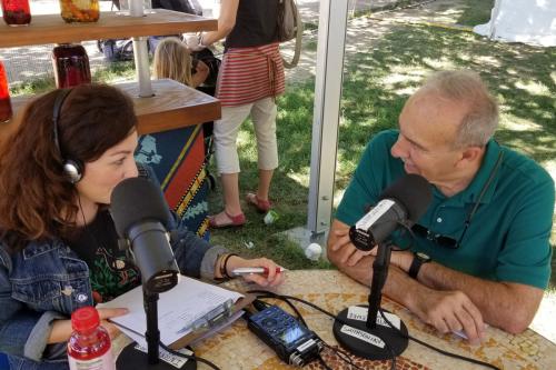 A man and a woman looking at each other sit at a table outside with recording equipment. Both people have a microphone in front of them that they are talking into.