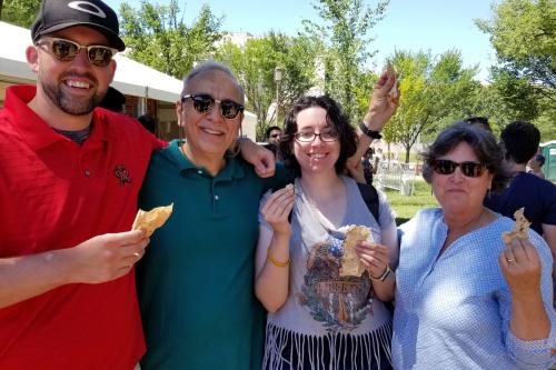 Two men and two women smile at the camera, holding up partially eaten pieces of bread. The four people are standing in front of several trees, with a few tents and chairs scattered behind them.