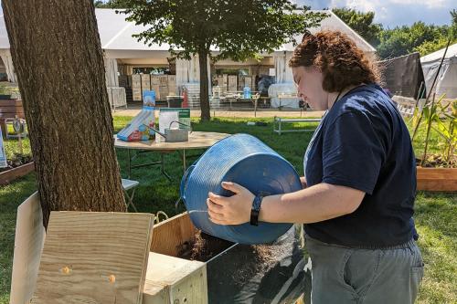 A young woman pours a bucket of soil into a raised wooden box with plexiglass on one side, making the interior visible.