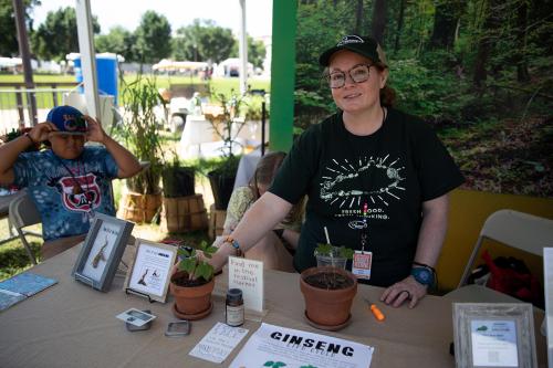 A woman stands at a booth outdoors, showing off a small green potted plant. A poster on the table reads Ginseng Life Cycle.