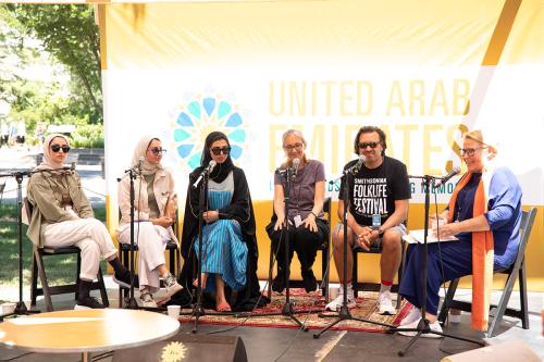 Six people on a small outdoor stage, each seated at a microphone.