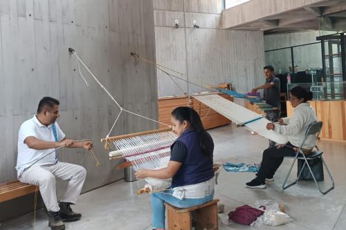 Four people work on backstrap looms attached to concrete walls.