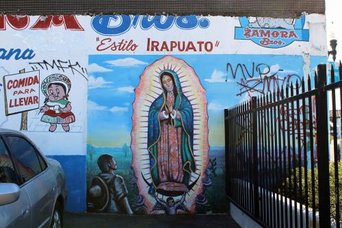 Painting of the virgin on the side of a commercial building, between a parked car and black iron fence. Next to her is a cartoon pig in green, white, and red with a sign that says in Spanish, Comida para llevar.