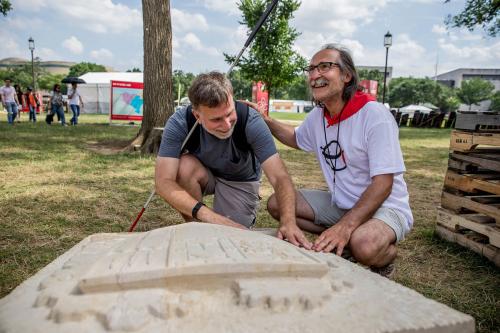 Two men crouch on the ground next to an off-white stone carving. One man holds his white cane under his arm and feels the carving with his hands.
