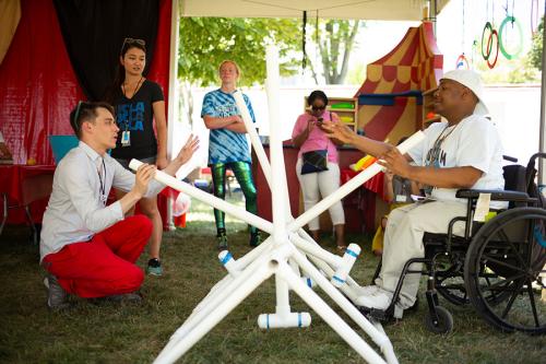 Two people sit on either side of a contraption made up white PVC pipe, each holding a pipe as a level. One of them uses a wheelchair, and the other crouches in the grass.