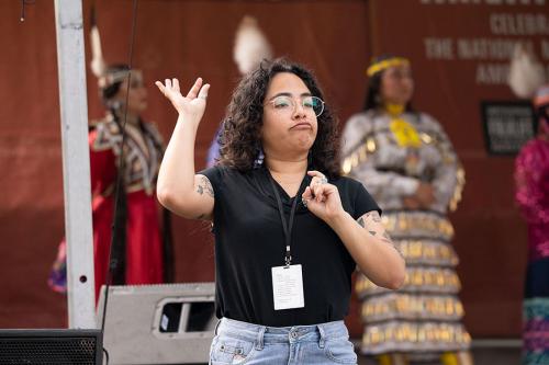 An ASL interpreter signs in front of people in Native regalia on stage.