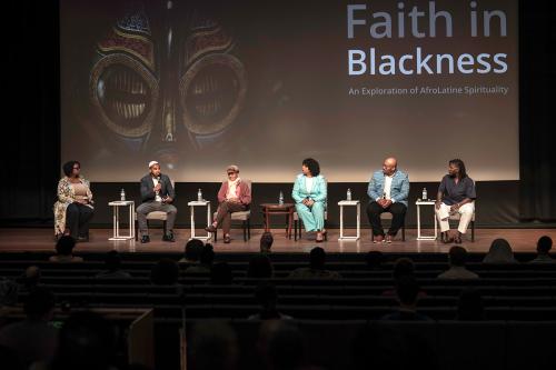 Six people seated on stage before a seated audience. On the projection screen behind them, a close-up on the eyes of a painted mask and the discussion title: Faith in Blackness: An Exploration of AfroLatine Spirituality.