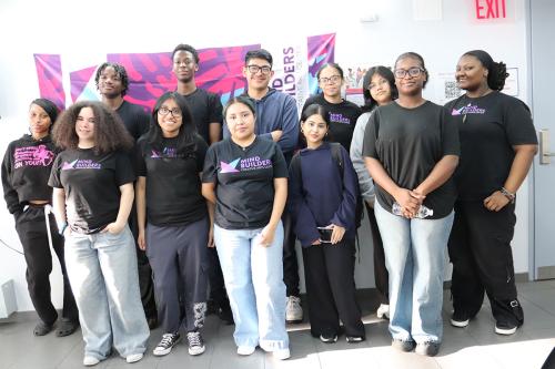 Twelve teens and young adults pose standing and smiling. Many wear black shirts with the pink, purple, and blue Mind Builders logo.