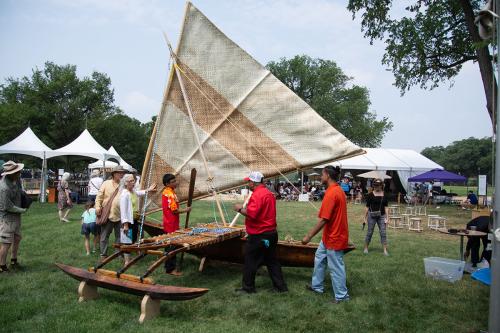 Three men in red shirts plus visitors stand around a wooden outrigger canoe set up on grass. The woven triangular sail is raised.