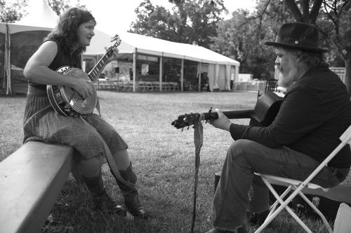 Facing each other, a woman plays banjo and a man plays acoustic guitar outdoors. Black-and-white photo.