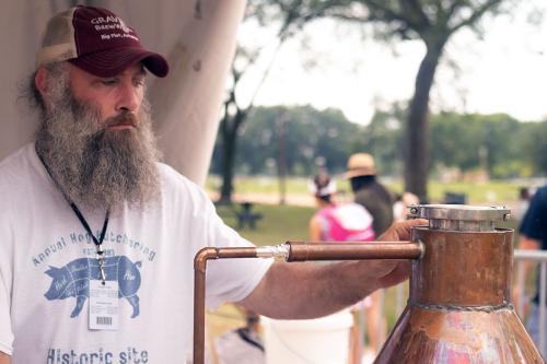 A man with a big gray beard and Annual Hog Butchering T-shirt inspects a copper still outdoors.