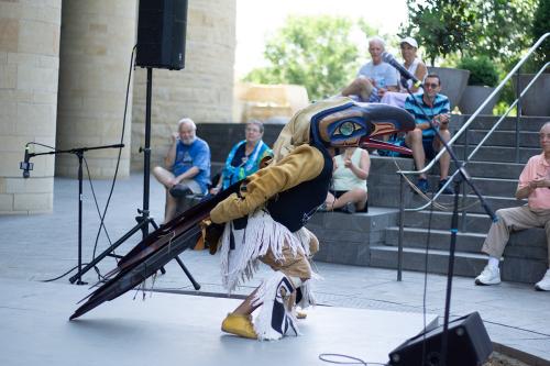 A man wearing birdlike regalia stands in front of an audience outdoors.