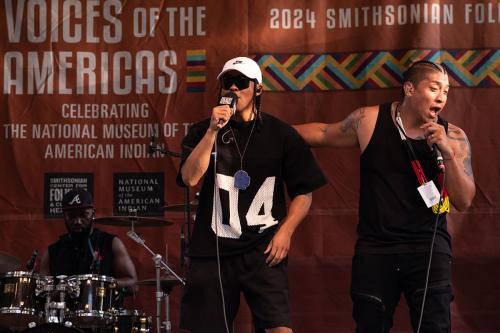 Two men in black clothing rap on stage, with a drummer on a drum kit playing in the background.